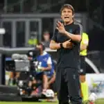 Antonio Conte, allenatore del Napoli, in panchina durante una partita, grida istruzioni ai suoi giocatori con espressione di intensa emozione e determinazione. Indossa una giacca nera e una sciarpa del Napoli, con il suo volto concentrato sulla partita. Sullo sfondo, lo stadio e i giocatori in azione sono sfocati.