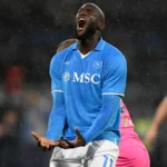 Romelu Lukaku, attaccante belga del Napoli, urla di frustrazione dopo aver sprecato un'occasione da gol durante una partita. Indossa la maglia del Napoli, con le braccia alzate in segno di frustrazione e un'espressione intensa di delusione sul volto. Sullo sfondo, il movimento del gioco e l'atmosfera dello stadio.