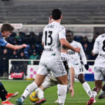 Charles De Ketelaere, calciatore belga dell'Atalanta, colpisce il pallone con il piede destro in un potente tiro in porta. La sua postura atletica e concentrata, con l'espressione determinata, cattura l'intensità del momento. Sullo sfondo, il campo di calcio sfocato con i giocatori avversari e la folla che incita