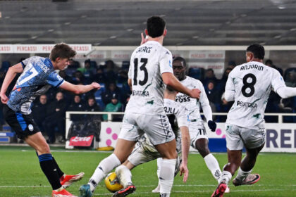 Charles De Ketelaere, calciatore belga dell'Atalanta, colpisce il pallone con il piede destro in un potente tiro in porta. La sua postura atletica e concentrata, con l'espressione determinata, cattura l'intensità del momento. Sullo sfondo, il campo di calcio sfocato con i giocatori avversari e la folla che incita
