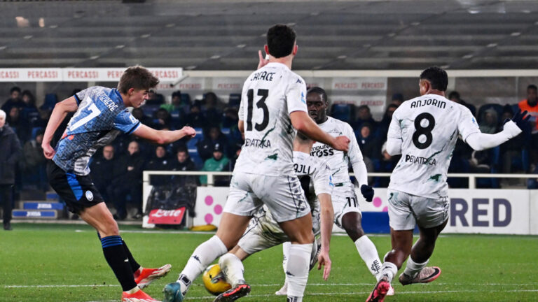 Charles De Ketelaere, calciatore belga dell'Atalanta, colpisce il pallone con il piede destro in un potente tiro in porta. La sua postura atletica e concentrata, con l'espressione determinata, cattura l'intensità del momento. Sullo sfondo, il campo di calcio sfocato con i giocatori avversari e la folla che incita